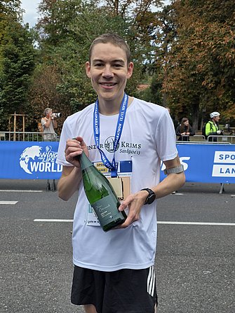 Das Foto zeigt Lukas Schneider, den zweitplatzierten Mann beim Viertelmarathon im Rahmen des Wachaumarathons 2026. Er trägt ein Winzer Krems Laufshirt und hält eine Magnumlflasche Sekt HAUS ÖSTERREICH von Winzer Krems in der Hand. 