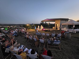 Open air Konzert im Weingarten der Winzer Krems von der Wiener Gruppe Stubenfliege. Das Bild zeigt den Blick vom Publikum aus auf die Bühne. Die Menschen sitzen entspannt in Liegestühlen. Das Bild wurde in der Abenddämmerung fotografiert und hat eine sehr angenehme, entspannte und stimmige Atmosphäre. Im Hintergund ist das Stift Göttweig zu sehen. 